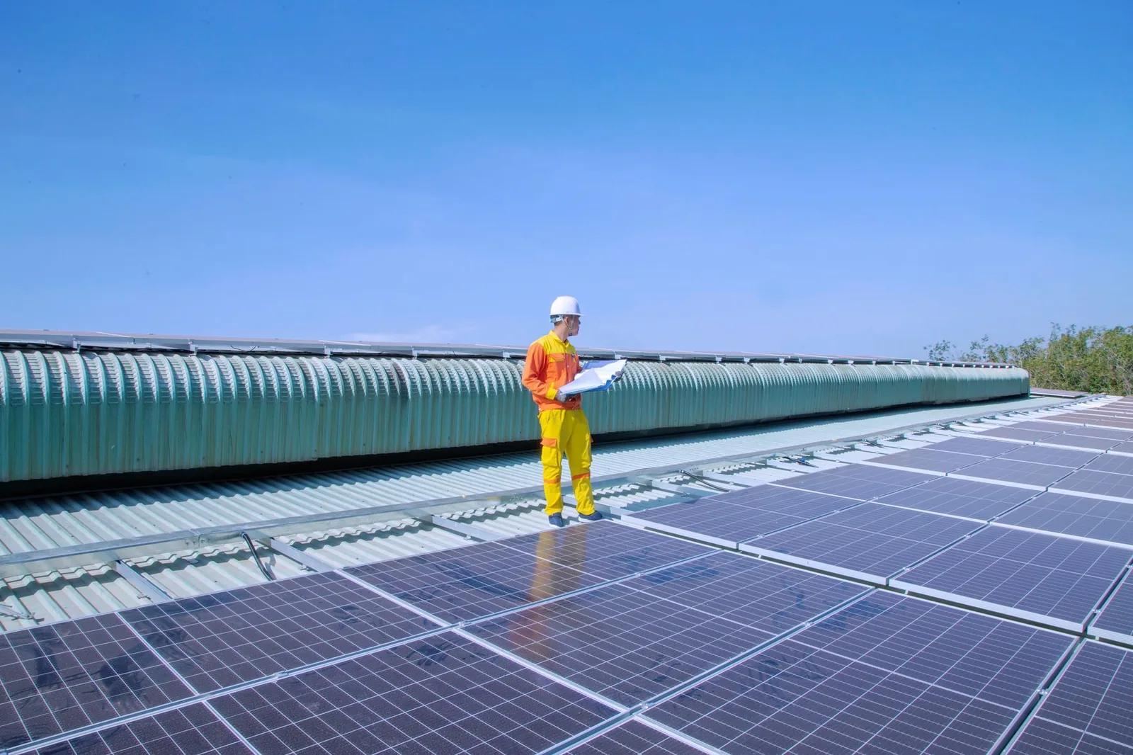 A technician in an orange and yellow uniform and white hard hat checking notes beside a commercial rooftop solar array